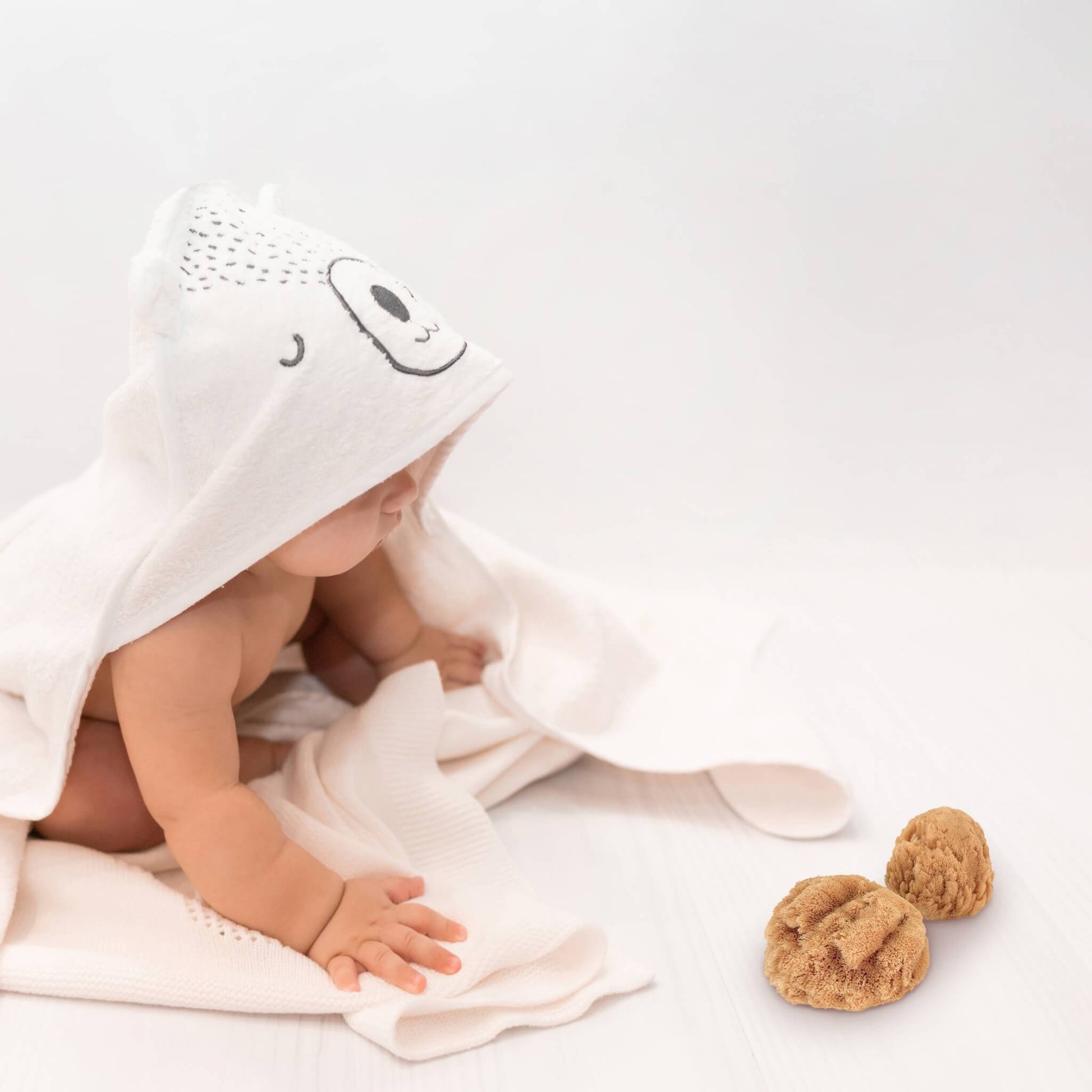 baby sitzt in weißem handtuch mit kapuze vor zwei naturschwämmen