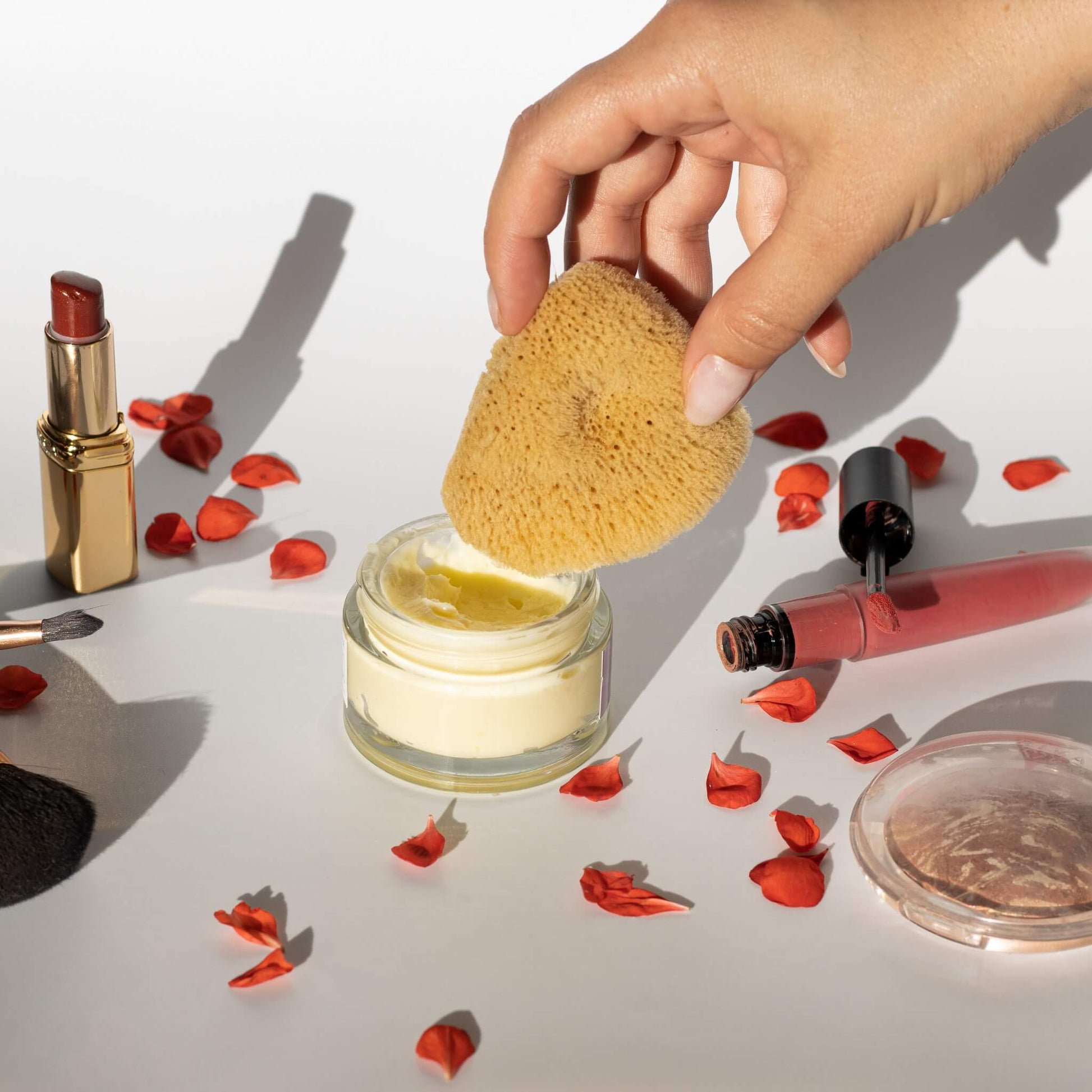 Gesichtsschwamm aus Naturschwamm wird aus einem Tiegel mit Creme entnommen, umgeben von Make-up und Rosenblättern