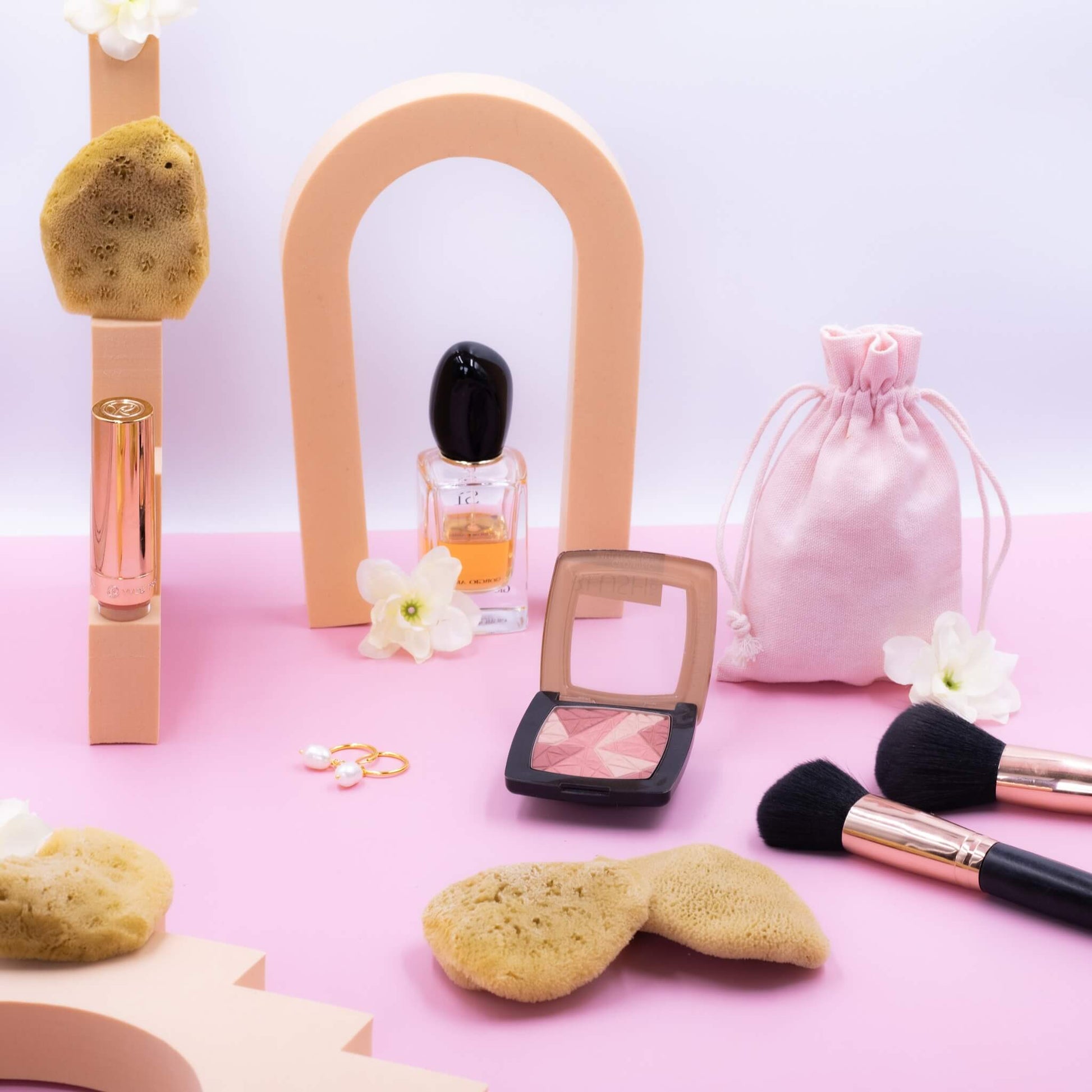 Drei Gesichtsschwämme aus Naturschwamm liegen auf rosa Untergrund zwischen Schminkpinseln, Rouge, Parfüm und einem rosa Beutel