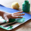 Eine Kinderhand drückt einen grün eingefärbten Malschwamm auf ein Papier als Teil eines kreativen Malprojekts, wobei die Hände farbig beschmiert sind.