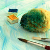 Ein mit blauer und gelber Farbe bedeckter Malschwamm liegt auf einem mit Aquarellfarben gemalten Hintergrund neben einem Pinsel und Farbtuben.