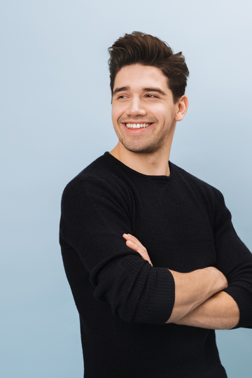 Junger Mann mit dunklem Haar und schwarzem Pullover, lächelnd mit verschränkten Armen vor einem blauen Hintergrund – Kunde von tom&pat Naturprodukte.