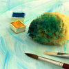Mit Wasserfarben eingefärbter Naturschwamm liegt neben Aquarellkasten und Pinseln – ideal als Malschwamm für Aquarelltechniken.