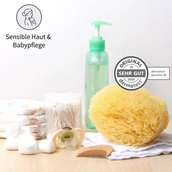 Baby Naturschwamm zur sanften Pflege sensibler Haut mit Dermatest-Siegel, umgeben von Babyartikeln wie Windeln, Wattepads und Pflegeflasche