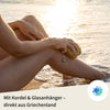 Person pflegt ihre Beine mit einem Naturschwamm am Strand bei Sonnenuntergang – ideal als Duschschwamm mit Kordel und Glashänger