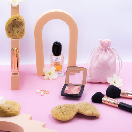 Drei Gesichtsschwämme aus Naturschwamm liegen auf rosa Untergrund zwischen Schminkpinseln, Rouge, Parfüm und einem rosa Beutel