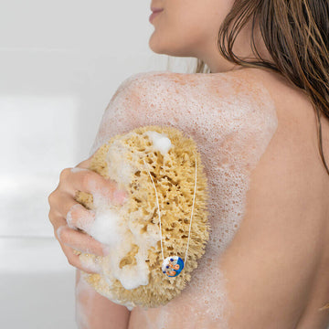 Frau reinigt sich unter der Dusche mit einem großen Naturschwamm – ideal für sanfte Körperpflege und Duschrituale